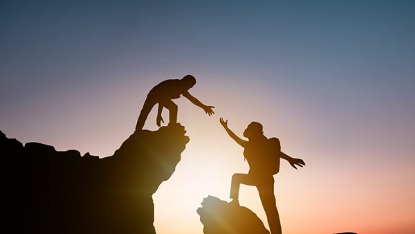 Together we can climb mountains. Person reaches for friend to help them climb upward.
